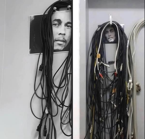 A creative yet messy blursed image showing two bundles of tangled black audio/visual cables hanging from wall hooks. Tape-mounted photos of Bob Marley’s face are positioned behind the hooks so the overflowing wires perfectly resemble his iconic dreadlocks.