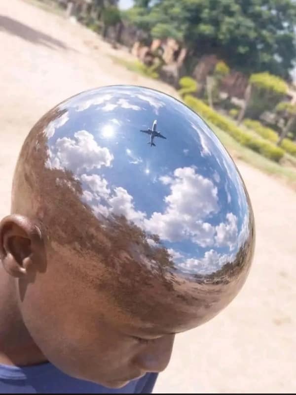 blursed image featuring the extreme close-up of a person's flawlessly bald, polished head. The surface is so reflective it acts as a mirror, showing a crystal-clear reflection of a blue sky, puffy white clouds, and a commercial jet flying directly overhead.