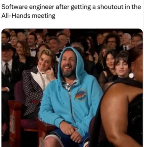 A classic meme capturing the pinnacle of tech culture. It shows actor Adam Sandler sitting in an awards ceremony crowd, wearing a casual bright blue zip-up hoodie while everyone around him is in formal black-tie attire. The caption reads: "Software engineer after getting a shoutout in the All-Hands meeting."