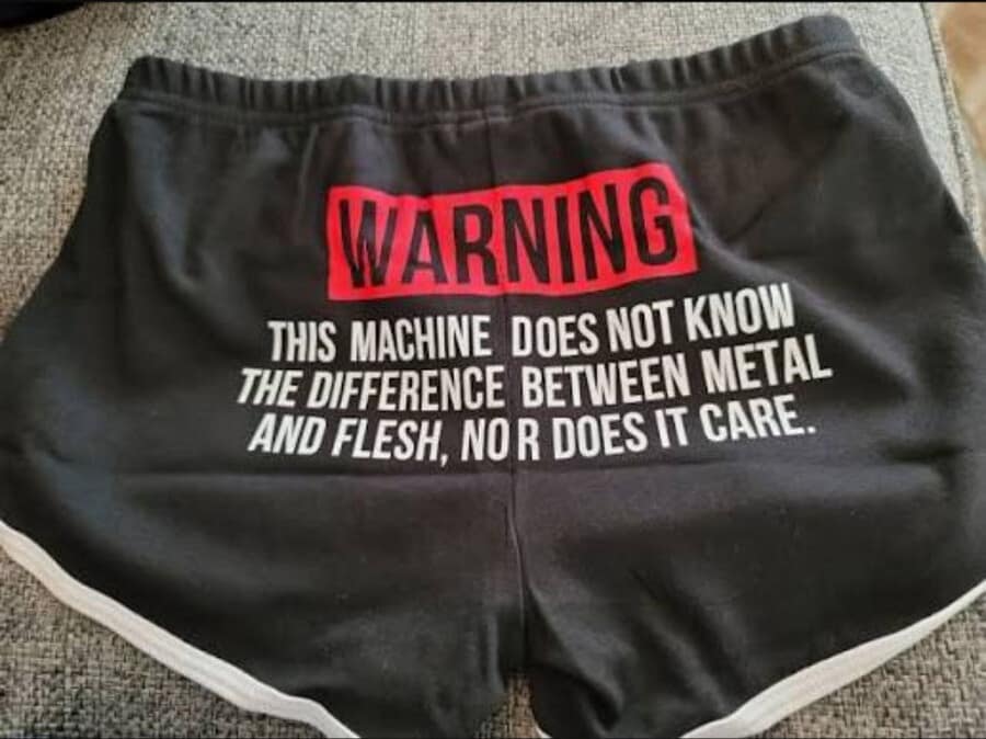 A bold and threatening classic meme showing the back of a pair of black gym shorts. A large red "WARNING" block is followed by the white text: "THIS MACHINE DOES NOT KNOW THE DIFFERENCE BETWEEN METAL AND FLESH, NOR DOES IT CARE."