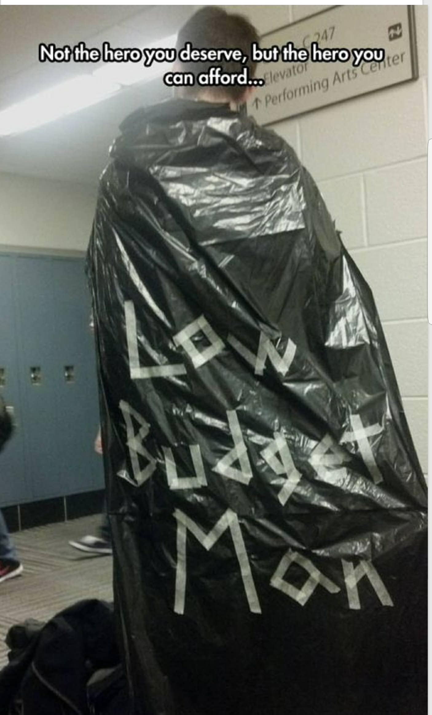 A low-budget superhero costume made from a black plastic trash bag. The words "LOW BUDGET MAN" are taped onto the back in white masking tape, accompanied by the text, "Not the hero you deserve, but the hero you can afford..."