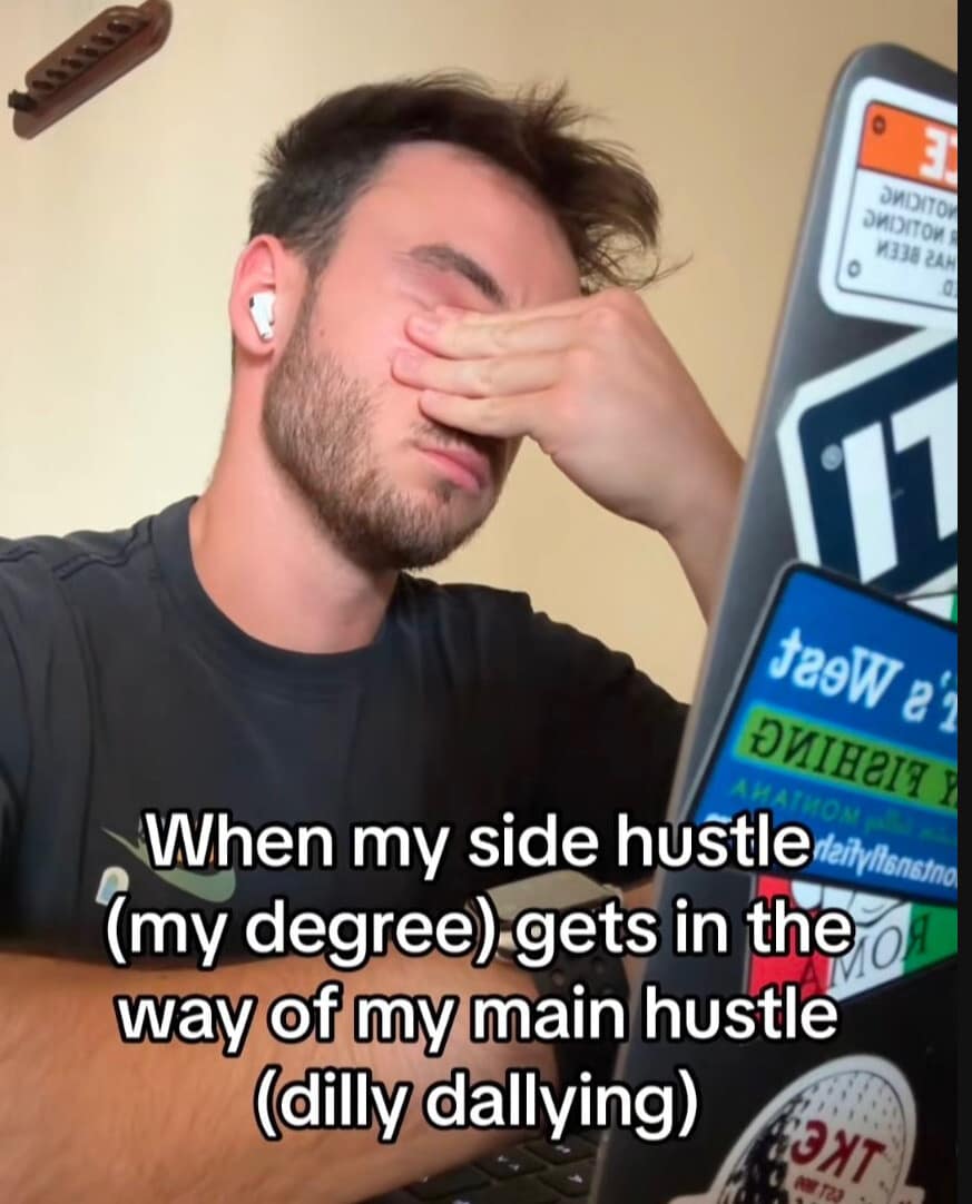 A man rubs his eyes in a state of academic burnout next to a laptop covered in stickers. The text describes a classic struggle: "When my side hustle (my degree) gets in the way of my main hustle (dilly dallying)."