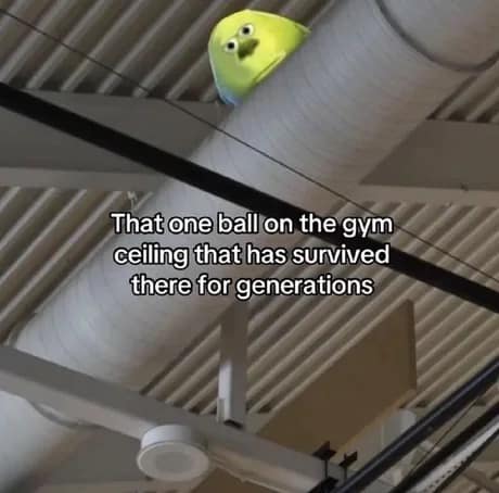 A classic meme looking up at a white industrial gym ceiling. Wedged on top of a large ventilation pipe is a yellow playground ball with Mike Wazowski's face photoshopped onto it, staring down with judgment. Text: "That one ball on the gym ceiling that has survived there for generations".