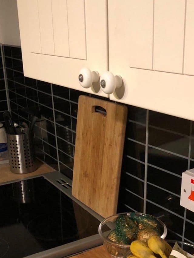 visual classic meme showing a "DIY security system" fail. A wooden cutting board has been perfectly wedged vertically between two white cabinet knobs, making it physically impossible to open the cupboard doors—a perfect example of over-engineering a simple problem.