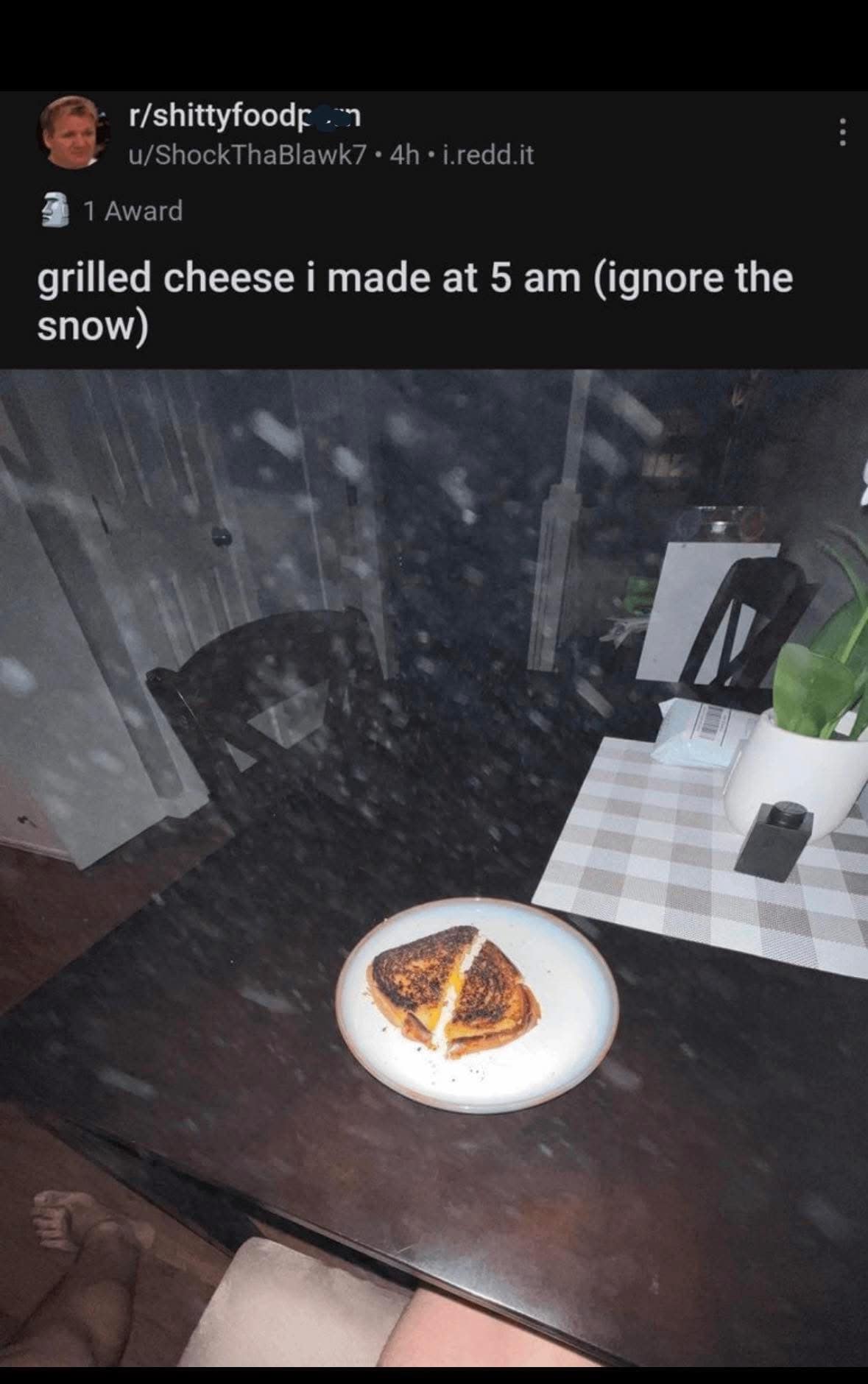 A chaotic entry from r/shittyfoodporn. A photo shows a charred grilled cheese sandwich on a table at 5 AM. The camera's flash catches a heavy cloud of floating dust particles, leading the poster to add the hilarious disclaimer: "(ignore the snow)."