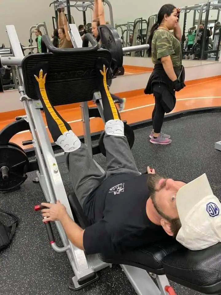 A man using a leg press machine at a public gym provides a jarring visual for this cursed image. He is wearing long, bright yellow socks that perfectly resemble scaly chicken legs, making it appear as though a giant bird is effortlessly pushing several hundred pounds of weight.