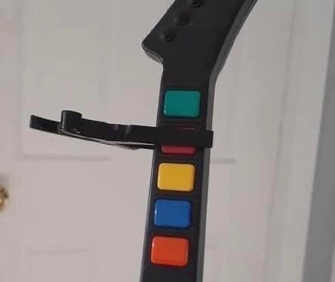 A cursed instrument photo showing a plastic Guitar Hero controller with a real guitar capo clamped firmly onto the neck over the colored fret buttons. This is inherently useless as the controller uses electronic switches rather than strings, making the "pitch change" purely decorative and frustrating.