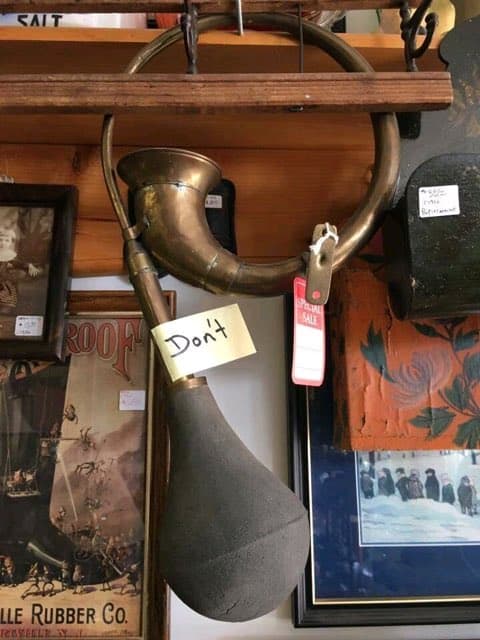 An ominous cursed instrument sighting in an antique shop. A vintage brass bulb horn hangs from a wooden shelf with a simple, handwritten yellow sticky note attached to the stem that just says: "Don’t."