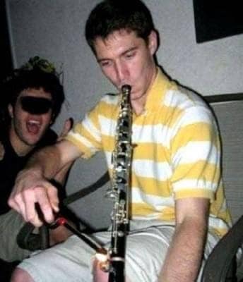 "cursed" grainy image of a man using a wooden clarinet as a makeshift smoking pipe. He is seen inhaling from the mouthpiece while holding a lighter to one of the lower tone holes, completely subverting the intended use of the woodwind instrument.
