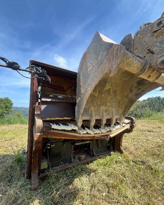 A dramatic and heavy cursed instrument photo of an old upright piano being crushed by the massive, dirt-caked bucket of an excavator. The keys are splintering and the internal strings are visible as the machinery provides a final, crushing "performance."