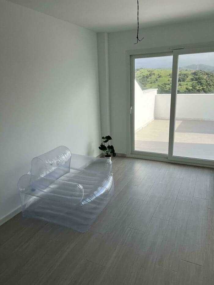 A minimalist design fail showing a stark white room with a single potted plant and a clear, plastic inflatable loveseat. It represents a peak example of temporary and questionable home decor in an otherwise empty apartment.