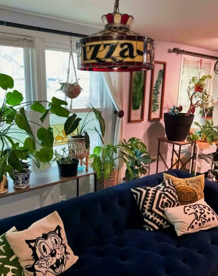 A cozy living room filled with houseplants, centered by a bizarre piece of questionable home decor: a large, vintage-style hanging lamp with "PIZZA" spelled out in stained glass, likely salvaged from a defunct 1980s-era pizza parlor.