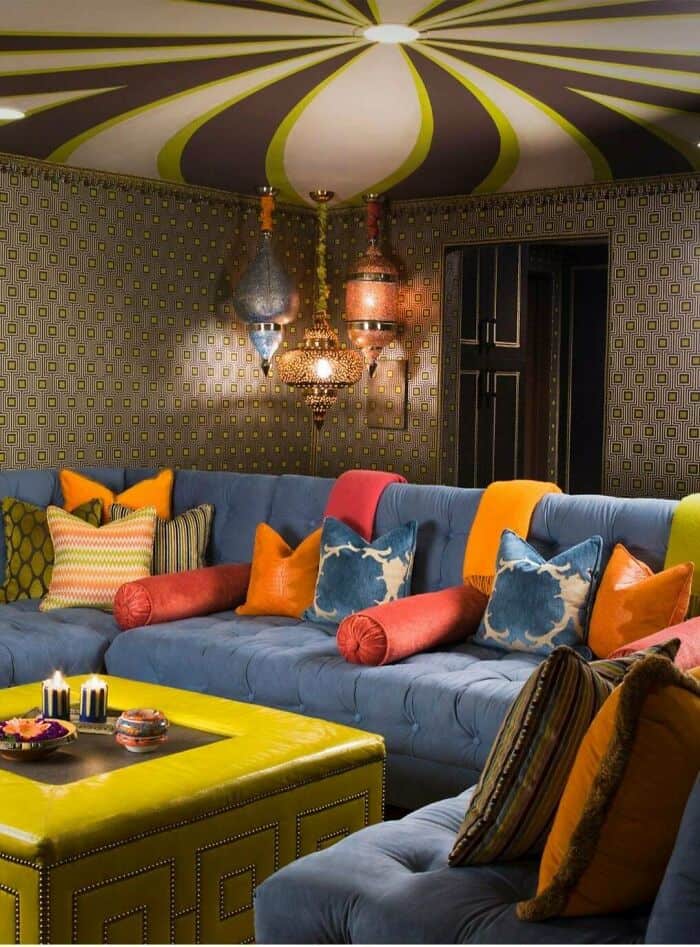 A visually chaotic living room defined by questionable home decor, including a yellow-and-purple striped "circus tent" ceiling, clashing patterned wallpaper, and a bright yellow ottoman paired with a massive blue tufted sofa.