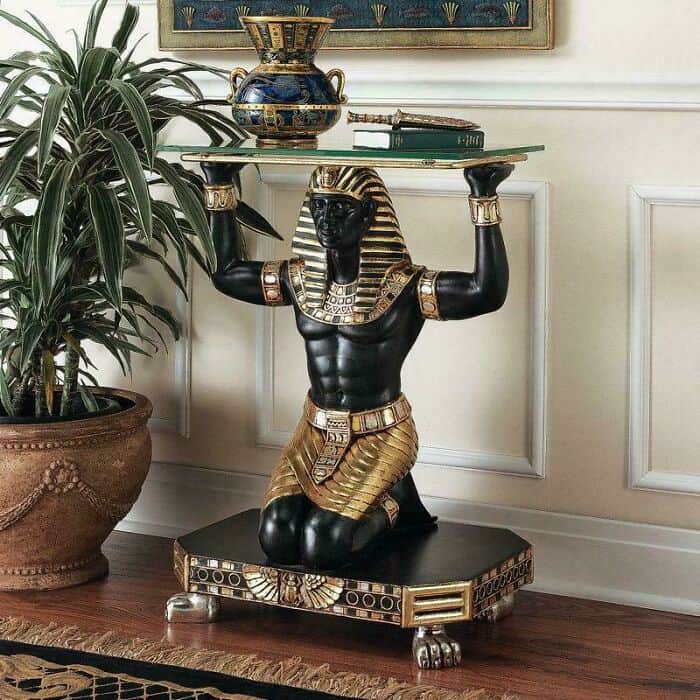 A piece of highly specific and questionable home decor consisting of a side table shaped like a kneeling Egyptian pharaoh figure, intricately detailed in black and gold, holding a glass pane over his head.
