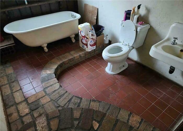 A disastrous bathroom design fail where an uneven, winding brick path has been laid directly into the middle of the tiled floor, creating a massive tripping hazard between the toilet and the clawfoot tub.
