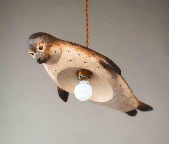 A bizarre lighting design fail consisting of a carved wooden seal hanging from the ceiling as a pendant lamp. A bare lightbulb protrudes directly from the animal's belly, creating a truly strange piece of questionable home decor.
