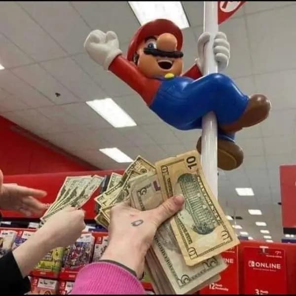 A chaotic dumb meme from inside a Target electronics section. A small Mario statue is shown sliding down a white display pole. Multiple hands are reaching into the frame, holding out $5 and $10 bills toward the statue as if they are tipping a performer.