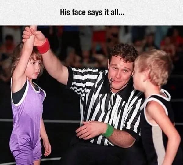 dumb meme featuring a youth wrestling referee with a look of pure, soul-shattering bewilderment. He is kneeling on the mat, holding up the arm of a tiny girl who has clearly just won, while staring at the defeated boy as if trying to process a glitch in the simulation.