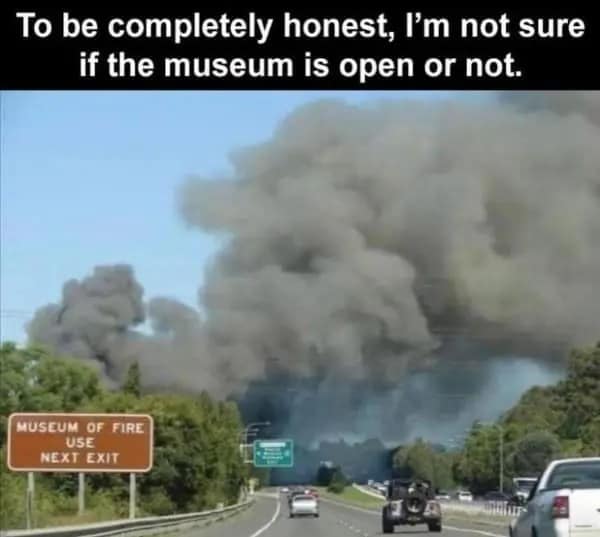 An ironic dumb meme taken from the perspective of a driver on a highway. A massive, towering plume of thick black smoke dominates the horizon. In the foreground, a brown road sign perfectly timed for the disaster reads: "MUSEUM OF FIRE USE NEXT EXIT."