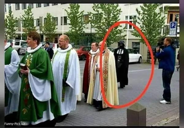 A hilarious dumb meme featuring a street procession of Catholic clergy in long robes and capes. Circled in a bold red ring in the background is someone in a full, screen-accurate Darth Vader costume, casually walking among the priests as if he’s part of the order.