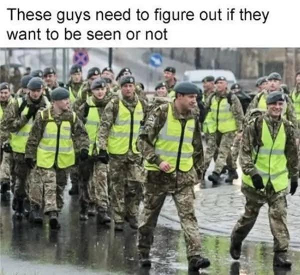 A logic-defying dumb meme showing a squad of soldiers marching in a city. While they are wearing full woodland camouflage uniforms intended for stealth, they are all also wearing neon yellow high-visibility safety vests, creating the world's most confused military aesthetic.