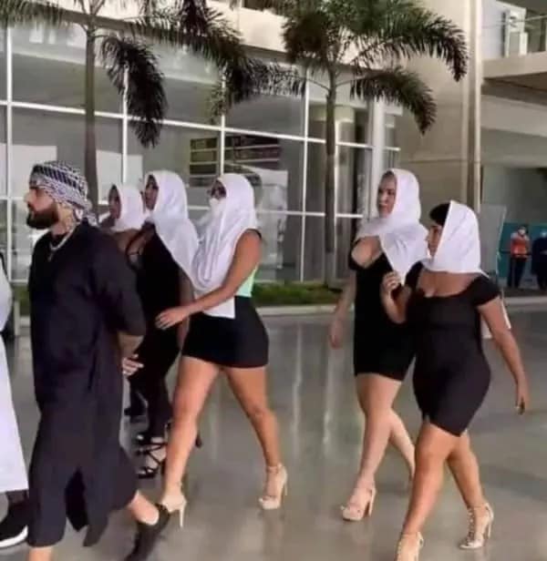 A bizarre dumb meme captured in a modern, glass-walled building. An Arab man in traditional black robes and a kaffiyeh leads a group of several women who are all wearing revealing, skin-tight black mini-dresses paired with white traditional headscarves.