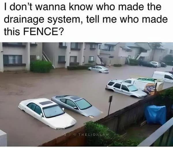 An engineering-themed dumb meme showing a massive flood in an apartment complex parking lot. While the cars are half-submerged, a simple brick and wood fence is somehow holding back several feet of water, creating a dry zone on the other side. The text asks: "I don't wanna know who made the drainage system, tell me who made this FENCE?"