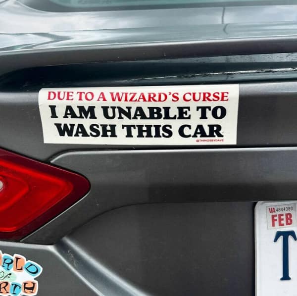 A funny bumper sticker on a grey car trunk that reads DUE TO A WIZARD'S CURSE I AM UNABLE TO WASH THIS CAR in red and black block text on a white background.