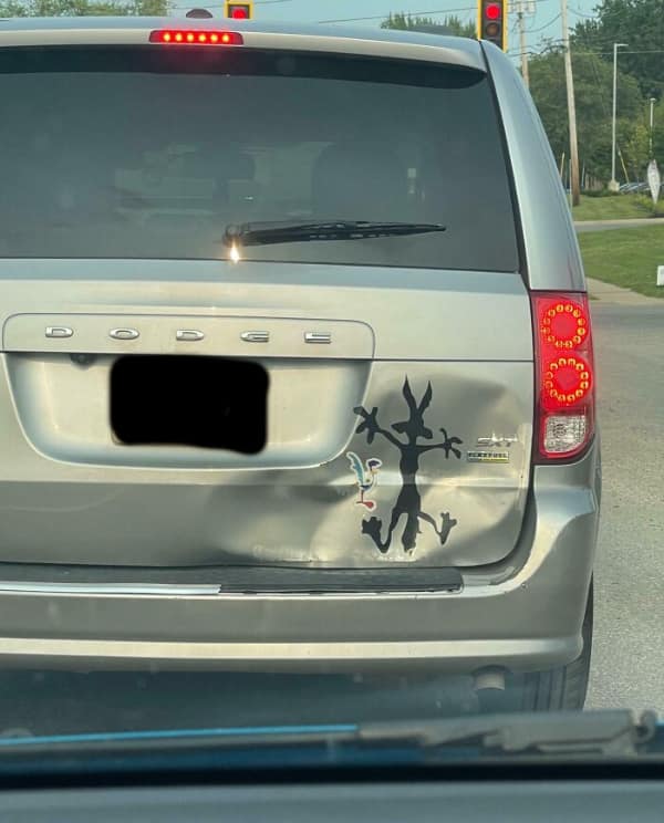 A rectangular [funny bumper sticker] on the silver bumper of a minivan, centered below the rear hatch. The text is printed in orange block letters on a black background, stating the driver's hard truth: "I USED TO BE COOL. NOW I DRIVE A MINIVAN."
