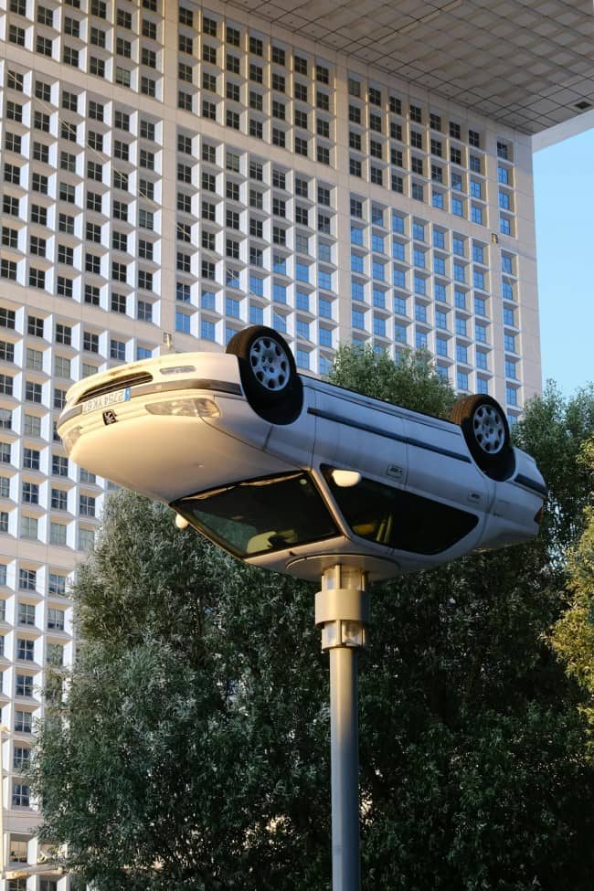 White sedan mounted upside down on top of urban street lamp post in front of large office building