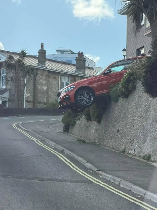 Orange red BMW perched at steep angle on top of stone retaining wall above road in coastal UK town