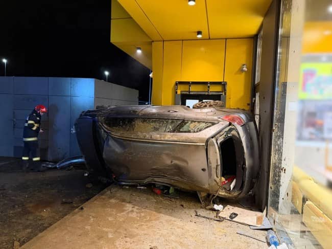 Heavily damaged car resting on its side inside yellow fast food restaurant drive-through at night with firefighter