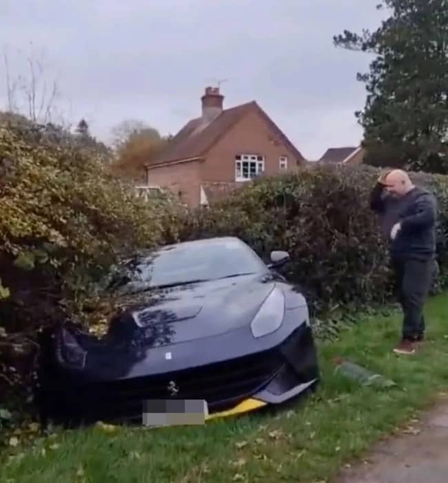 Dark Ferrari sports car crashed nose-first into roadside hedge while distressed bald man stands nearby