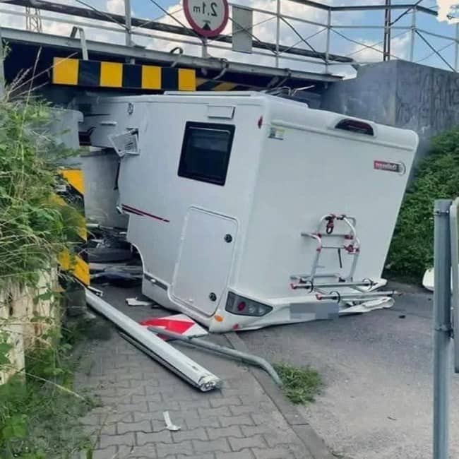 Silver car driven off road into ditch at sharp angle beside road sign with person's legs visible standing nearby