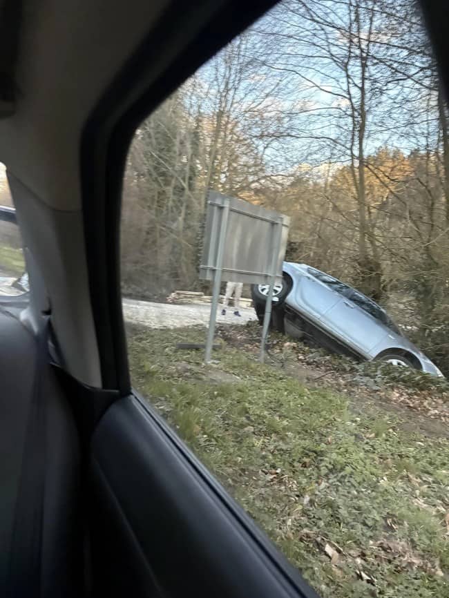 Silver car driven off road into ditch at sharp angle beside road sign with person's legs visible standing nearby