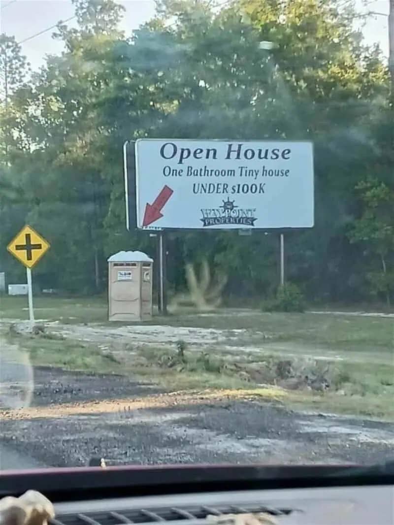 Real estate billboard for a tiny house pointing directly at a portable toilet outdoors.