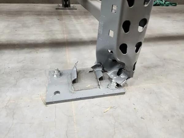 A bone-chilling funny picture of a warehouse pallet rack upright that has been crushed into a twisted metal mess at the base, yet continues to hold up the entire shelf.