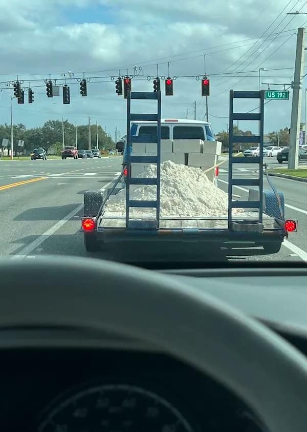 A "highway hazard" funny picture showing a trailer carrying an uncovered, unsecured mountain of white sand and concrete blocks through a busy intersection.