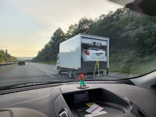 A "Tetris fail" funny osha violation picture showing a white SUV stuffed into the back of a box truck that is clearly too small, with the liftgate wide open and the car secured only by a few yellow ratchet straps tied to the bumper while flying down the highway.