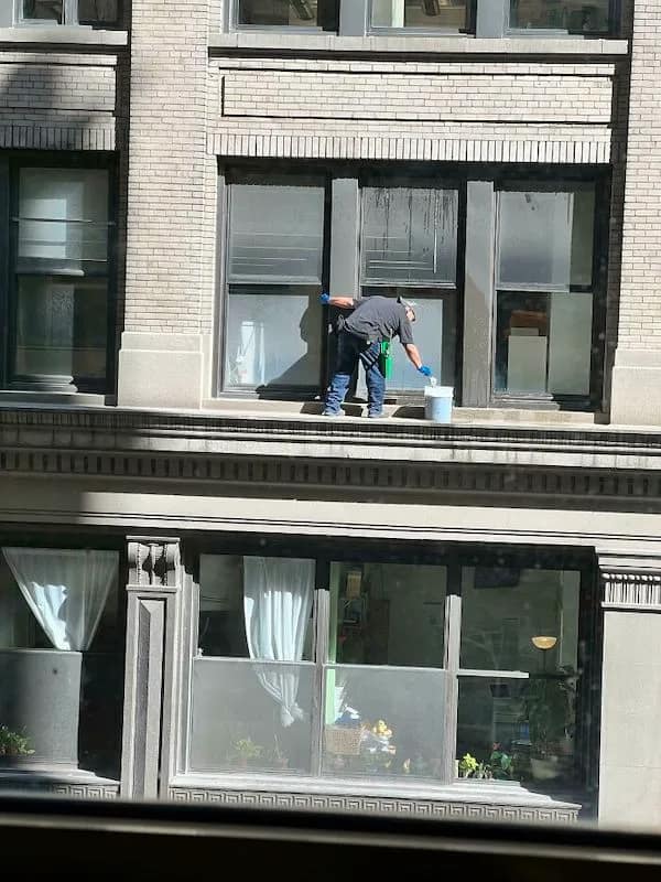 A terrifyingly funny osha violation picture of a window washer standing on a three-inch decorative ledge several stories up, leaning back to dip his brush into a bucket without a single harness, rope, or shred of self-preservation in sight.