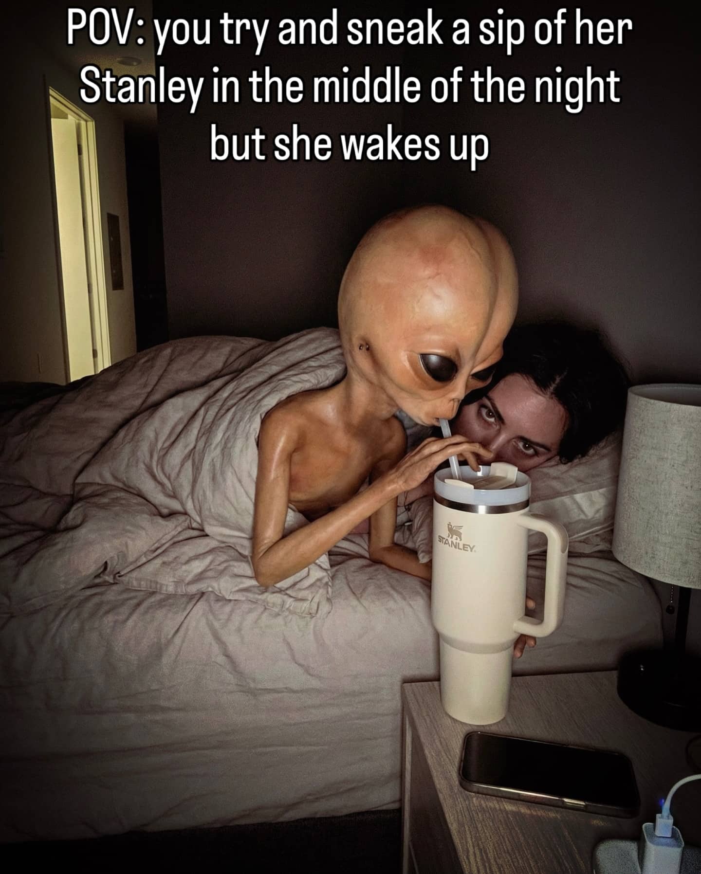 Alien drinking from a Stanley cup at night while a woman watches from the bed.