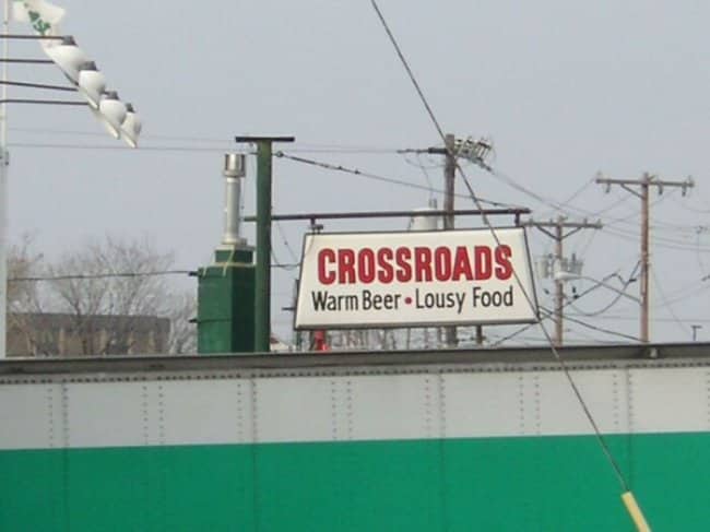 Crossroads bar sign honestly advertising "Warm Beer • Lousy Food" with refreshing brutal transparency.