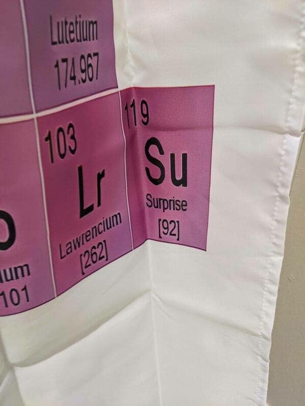 A scientific funny shower curtain featuring a detailed Periodic Table of Elements. However, tucked into a corner is a fake element with the symbol "Su," the name "Surprise," and the atomic number 119, designed to catch the eye of unsuspecting chemists.