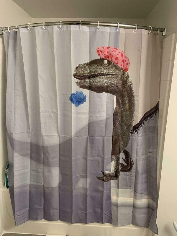 A prehistoric funny shower curtain showcasing a realistic velociraptor that has been edited to wear a bright pink polka-dot shower cap. The dinosaur is shown clutching a blue loofah, ready for its morning exfoliation.