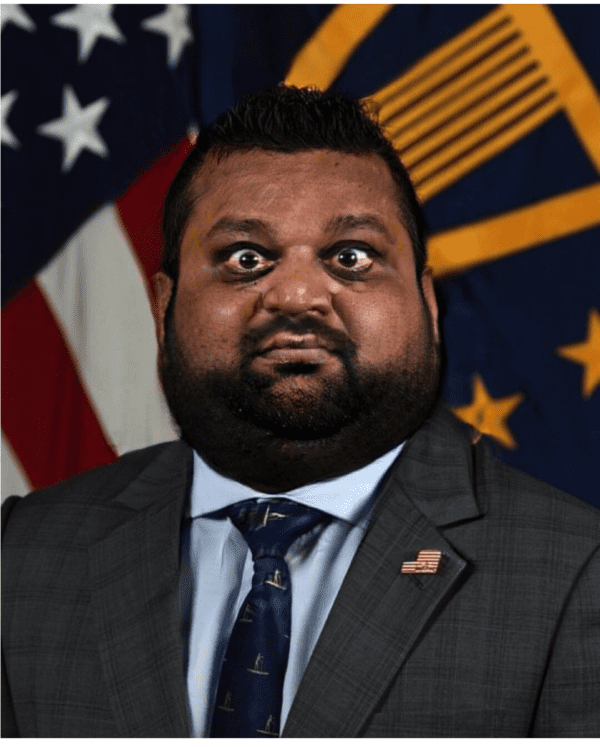 A surreal, photoshopped Kash Patel meme where his features are compressed and widened. He appears in a grey suit and American flag pin, but his head is flattened and his eyes are shrunk into a state of permanent, wide-eyed bewilderment.
