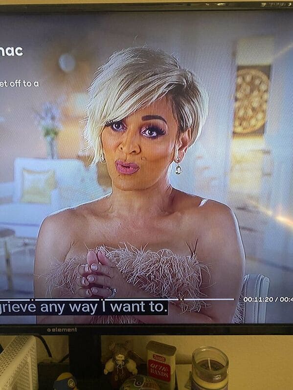 A screenshot from a reality show capturing a makeup fail with jaundiced, yellow-toned foundation. The look is finished with frosted pink lips and heavy, unblended purple eyeshadow that emphasizes the under-eye area.