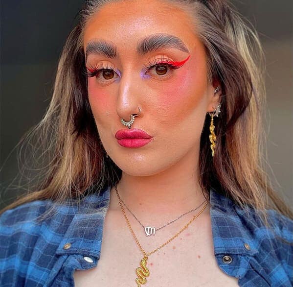 A vibrant makeup fail featuring a woman with an overly saturated orange complexion. The look includes thick, dark eyebrows, neon red winged eyeliner, and bright magenta lipstick that creates a high-clash aesthetic against her blue flannel shirt.
