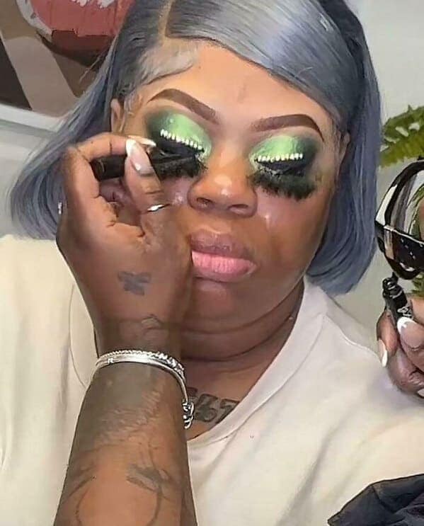 A high-intensity makeup fail showing a person with blue hair wearing a thick application of neon green eyeshadow. Small white decorative beads line the crease, while unblended yellow concealer and extremely heavy, drooping false lashes dominate the eye area.