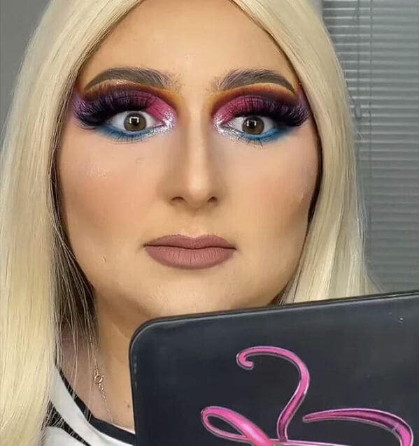 A startling makeup fail with wide-eyed intensity. The eye makeup consists of a chaotic blend of purple, pink, and blue glitter, topped with dark, uneven eyebrows and a matte nude lip that nearly disappears into the heavy foundation.