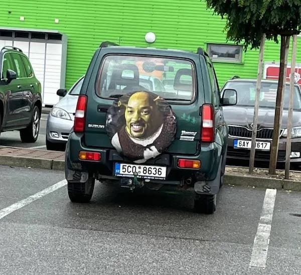 A hilarious funny meme photo of the back of a green Suzuki Jimny SUV parked in a lot. The spare tire cover features a cursed mashup of Will Smith's face and Shrek's green skin and ears, creating a Will-Shrek icon.